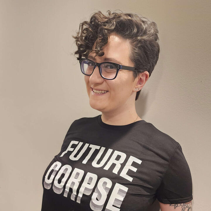 nonbinary white person with short curly brown hair and black thick framed glasses standing in front of a white wall wearing a black tshirt that says FUTURE CORPSE in white lettering